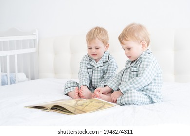 Two Toddler Baby Twin Boys In Pajamas Reading Book Sitting On White Bedding On Bed