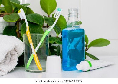 Two Toothbrushes In A Glass Cup Mouthwash A Tube Of Toothpaste And A Rolled Up Towel On The Dressing Table Against A Backdrop Of Green Foliage. High Quality Photo