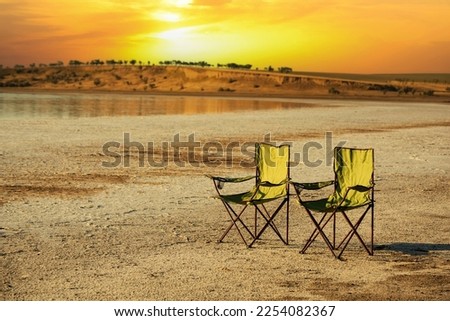 Two tourist chairs in a desert area on a lake at sunset. Secluded vacation in the wild.