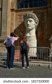 two tourists contemplate the head of the emperor Caesar Octavian Augustus on a pedestal illuminated from the side by the sun