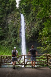 Two Tourists Looking At The La Fortuna Waterfall In Costa Rica. The Waterfall Is Located On The Arenal River At The Base Of The Dormant Chato Volcano.