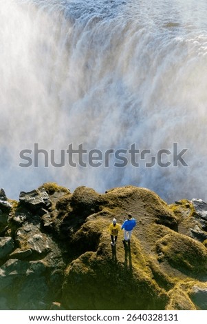 Two tourists stand at the edge of a rocky cliff, gazing at the breathtaking Dettifoss waterfall in Iceland.