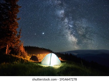 Two tourists tents, one of which shining white light on mountain grass hill with view on Milky Way galaxy. Night forest and peaks other mountain hills on the background. - Powered by Shutterstock