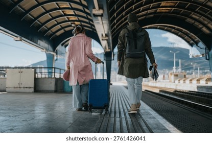 Two tourists walk down a covered train platform, one pulling a blue suitcase. A couple heads toward the tracks with backpacks and travel documents in hand.