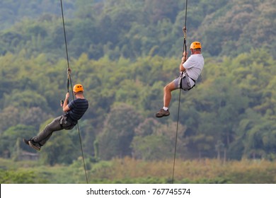 Two Tourists Wearing Casual Clothing Are Playing Zip Line And Selfie With Big Mountain Background At Chiang Rai, Thailand