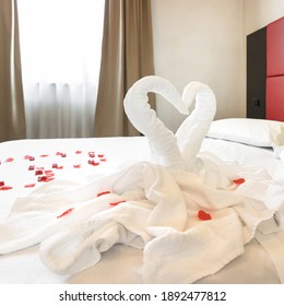 Two Towel Swans And Rose Petals On Bed In Light Hotel Room. For Couple In Honey Moon And Valentine's Day.