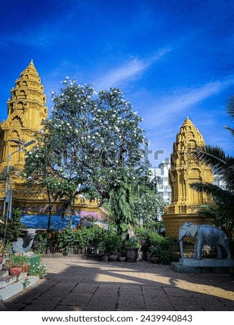 Two towering golden pagodas stand at the entrance of a sacred temple complex, flanked by lush, green trees and vegetation, against a backdrop of a vivid blue sky.Phnom Penh, Cambodia, 21.12.22