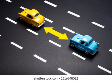 Two toy cars on the road one after another. One is blue one is yellow colored.