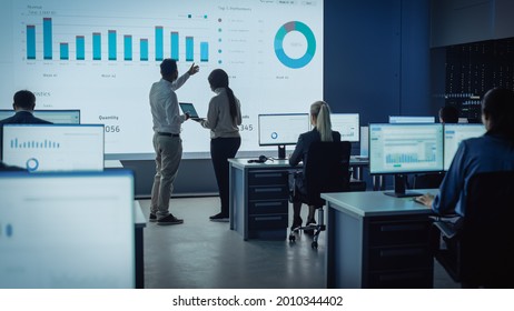 Two Traders Having a Meeting in a Modern Monitoring Office with Analytics Feed on a Big Digital Screen. Monitoring Room with Brokers and Finance Specialists Sit in Front of Computers. Colleagues Talk. - Powered by Shutterstock
