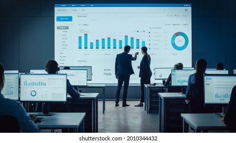 Two Traders Having a Meeting in a Modern Monitoring Office with Analytics Feed on a Big Digital Screen. Monitoring Room with Brokers and Finance Specialists Sit in Front of Computers. Colleagues Talk. - Powered by Shutterstock