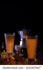 Two Transparent Glasses With Latte, A Coffee Maker In The Background On A Wooden Table With Chocolate, Anise, Hazelnuts, Lit By A Candle Flame. Black Background.