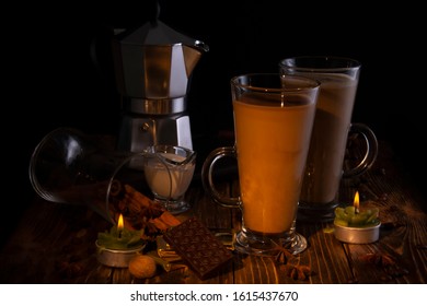 Two Transparent Glasses With Latte, A Coffee Maker In The Background On A Wooden Table With Chocolate, Anise, Hazelnuts, Lit By A Candle Flame. Black Background.