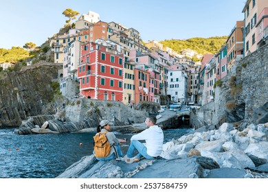 Two travelers relax on the rocky shore, surrounded by the vibrant buildings of Riomaggiore in Cinque Terre, Italy, a diverse couple of man and woman sitting by the ocean - Powered by Shutterstock