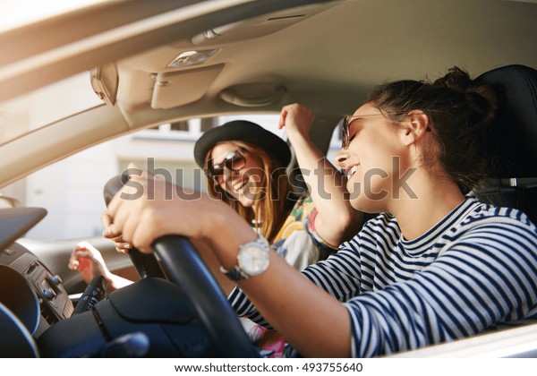 Two trendy attractive young woman singing along to\
the music as they drive along in the car through town viewed\
through the open side\
window