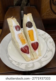 Two triangular fruit sando sandwiches filled with whipped cream, grapes, orange slices, and strawberries on a decorative white plate