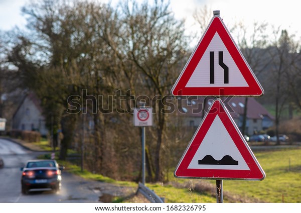 Two triangular red and white\
warning signs. The upper sign warns of a bottleneck. The lower sign\
warns of an uneven road or ruts. A car brakes in the\
background.