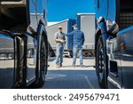 Two truck drivers engage in a conversation about logistics while standing between parked trucks in a transport yard. The bright blue sky adds to the vibrant atmosphere of this bustling environment.