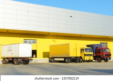 Two truck trailers and two trucks parked in front of a yellow factory. There is one red truck, one yellow truck, one yellow trailer and one white trailer. The factory door is open. - Shutterstock ID 1357002119
