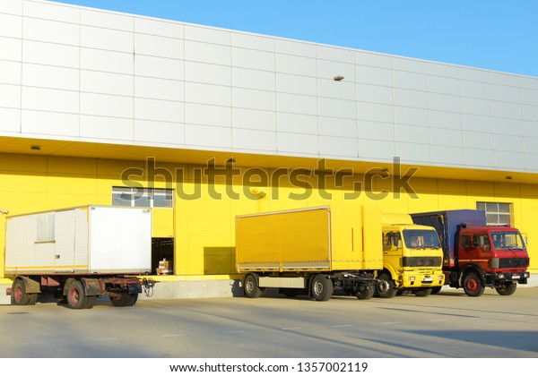 Two truck\
trailers and two trucks parked in front of a yellow factory. There\
is one red truck, one yellow truck, one yellow trailer and one\
white trailer. The factory door is\
open.