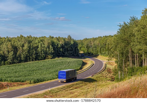 Two trucks are moving\
towards each other along a bend in the road in a rural area, rural\
field, trucks driving on the road near by agricultural fields and\
forest.