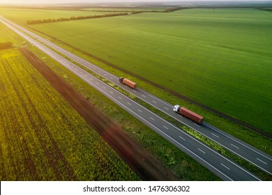 Two Trucks On The Higthway Sunset. Cargo Delivery Driving On Asphalt Road Along The Green Fields. Seen From The Air. Aerial View Landscape. Drone Photography.