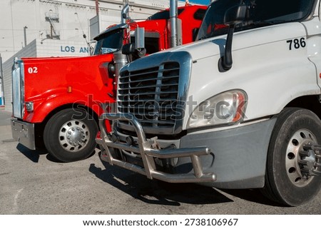 Two trucks are parked side by side in a Los Angeles area. One truck is red, while the other is white. The vehicles are positioned on a paved surface.