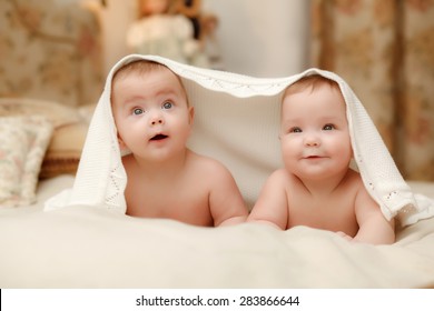 Two twin babies, seven-month  smiling girls covered with a white towel in bed on white sheets
