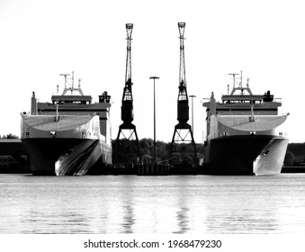 Two twin boats sit in dock next to cranes.