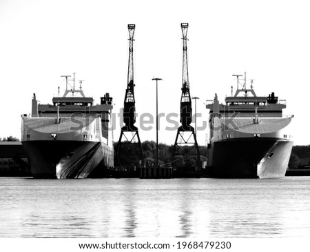Two twin boats sit in dock next to cranes.