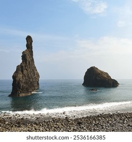 Two unique rock formations rise from the ocean near a rocky shoreline under a clear sky. Dramatic sea stacks add a rugged charm to the tranquil coastal scenery, with gentle waves lapping at the shore. - Powered by Shutterstock