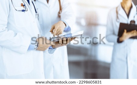 Two unknown doctors with stethoscopes discussing medical exam resoults, standing at sunny hospital office. Physicians use a computer tablet for filling up medication names records.