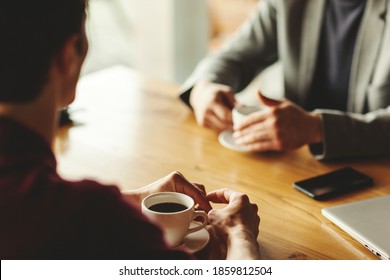 Two unrecognizable businessmen having business talk over coffee sitting at table in cafe. Laptop, coffee cups and smartphone on table. Focus on hands and cup of espresso - Powered by Shutterstock