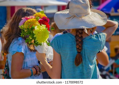 Two Unrecognizable Girls At Outdoor Farmers Market - On With Back To Camera Hold Boquet Of Flowers Hiding Others Face - She Has A Hat And A Pigtail And Is Holding Money In Her Hand - Bright And Colorf