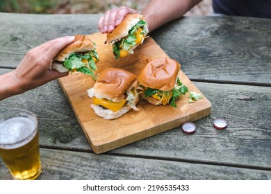 Two Unrecognizable Hands Pick Up Cheeseburgers From A Wooden Plate. Suburban Lifestyle.