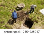 Two unrecognizable men are working in a field, one of them is using a shovel to dig a hole. The other man is holding a wheelbarrow, ready to transport the dirt. The scene is peaceful and quiet