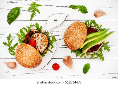 Two Veggies Burgers Served On Cutting Board Over White Rustic Table. Vegan Grilled Eggplant, Arugula, Sprouts And Pesto Burger. Veggie Beet And Quinoa Burger With Avocado. Top View, Overhead, Flat Lay