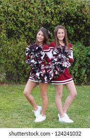 Two very cute teen aged cheerleaders posing outdoors on a green lawn.