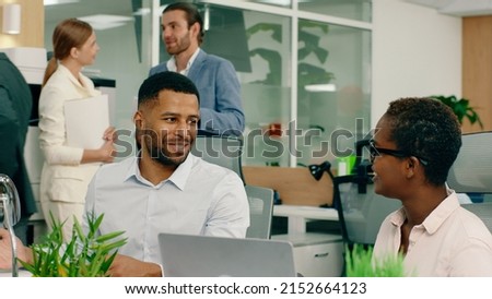 Two very good looking co- workers are in the office, having a conversation and giving each other good advice and ideas for work, a man is at a printer in the back.