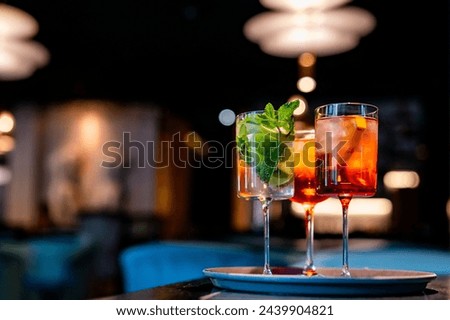 Two vibrant cocktails adorned with mint leaves rest on a tray in an elegant, softly lit bar setting. One is a green mojito, the other a reddish-orange concoction, both served in long-stemmed glasses
