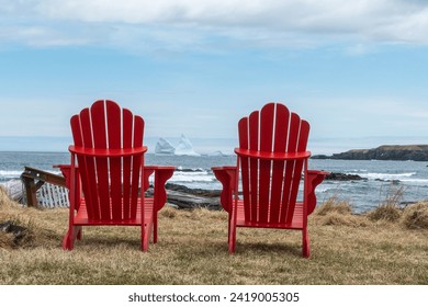 Two vibrant red empty Adirondack chairs face the Atlantic Ocean and cloudy blue sky on a grassy coastline. There's a large white glacier iceberg floating in the distance between the two chairs.   