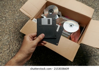 Two vintage floppy disks held in hand in front of a cardboard box full of old cd's and storage devices