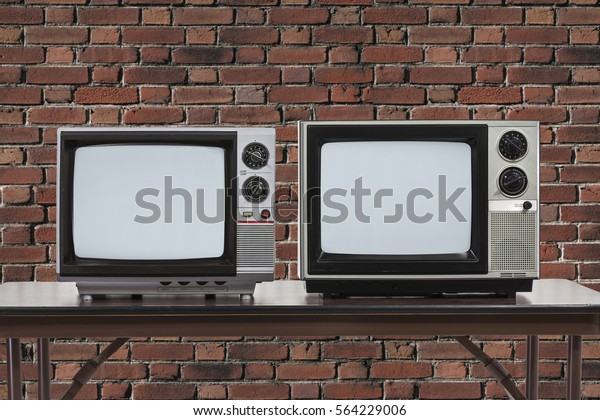 Two vintage\
televisions on table with brick\
wall.