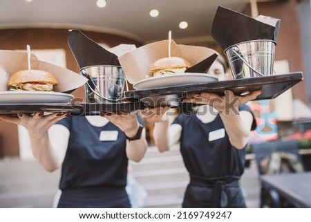 Two waiters serve table, take out burgers to the outdoor summer area in a cafe for a large company. Street fast food. Hands and tray close-up, no face. image with selective focus and noise effect