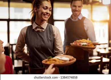Two waiters serving lunch and brining food to their gusts in a tavern. Focus is on happy waitress. 