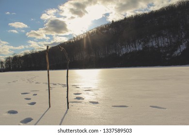 Two Walking Sticks Stuck Into The Ice On A Frozen Lake With Two Footprint Paths Coming Together. The Sun Is Setting Over The Mountains In The Background In A New England Forest In Winter. Fresh Snow