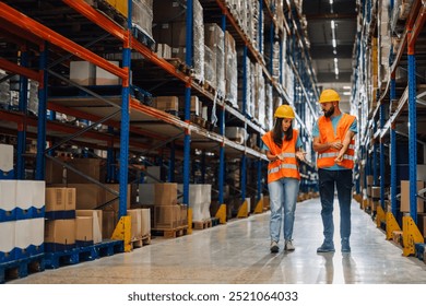 Two warehouse employees in safety gear walk through an industrial aisle while talking. They analyze and coordinate tasks related to inventory management in the stockroom. - Powered by Shutterstock