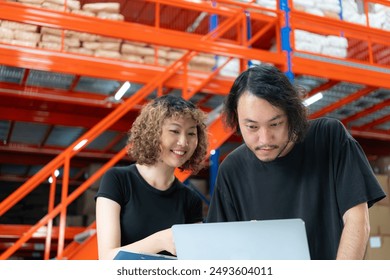 Two warehouse workers collaborating on a laptop in a warehouse setting, surrounded by shelves of packed goods and industrial equipment. - Powered by Shutterstock