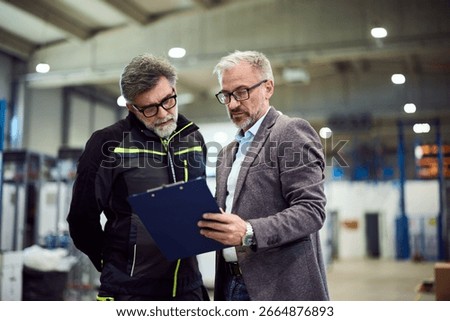 Two warehouse workers collaborating over a clipboard document inside an industrial building.