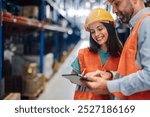 Two warehouse workers smiling and collaborating with a digital tablet in a busy logistics center. Surrounding by shelves and forklifts, they manage inventory efficiently