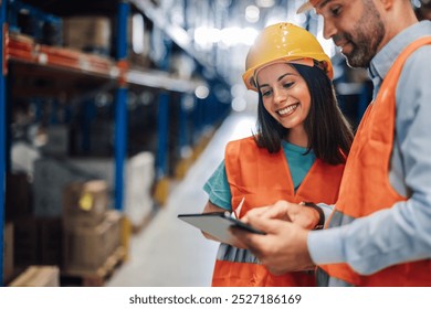 Two warehouse workers smiling and collaborating with a digital tablet in a busy logistics center. Surrounding by shelves and forklifts, they manage inventory efficiently - Powered by Shutterstock